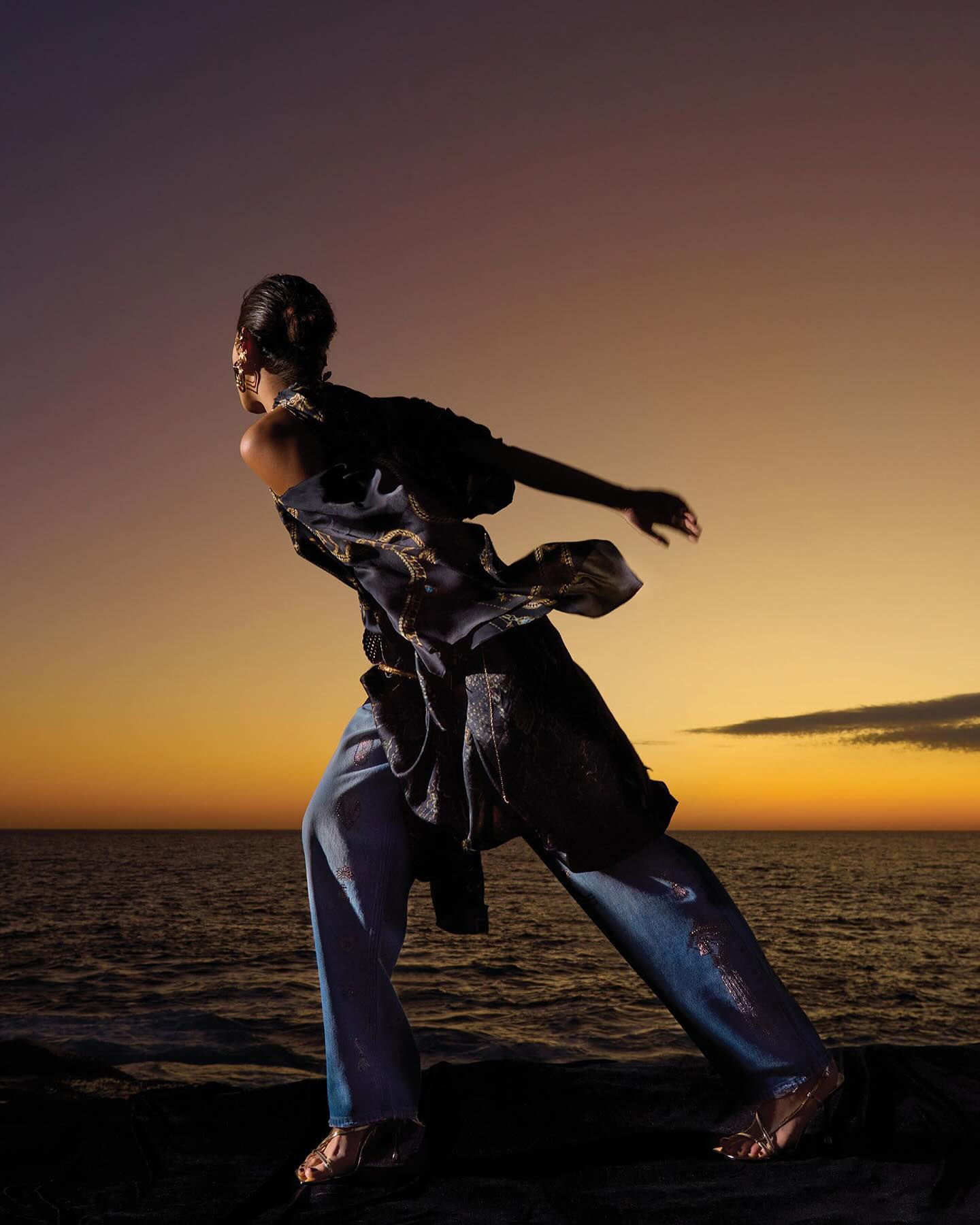 Woman in draped top and wide-leg jeans in a dynamic pose on rocks at sunset by the ocean, evoking a sense of movement and fashion.
