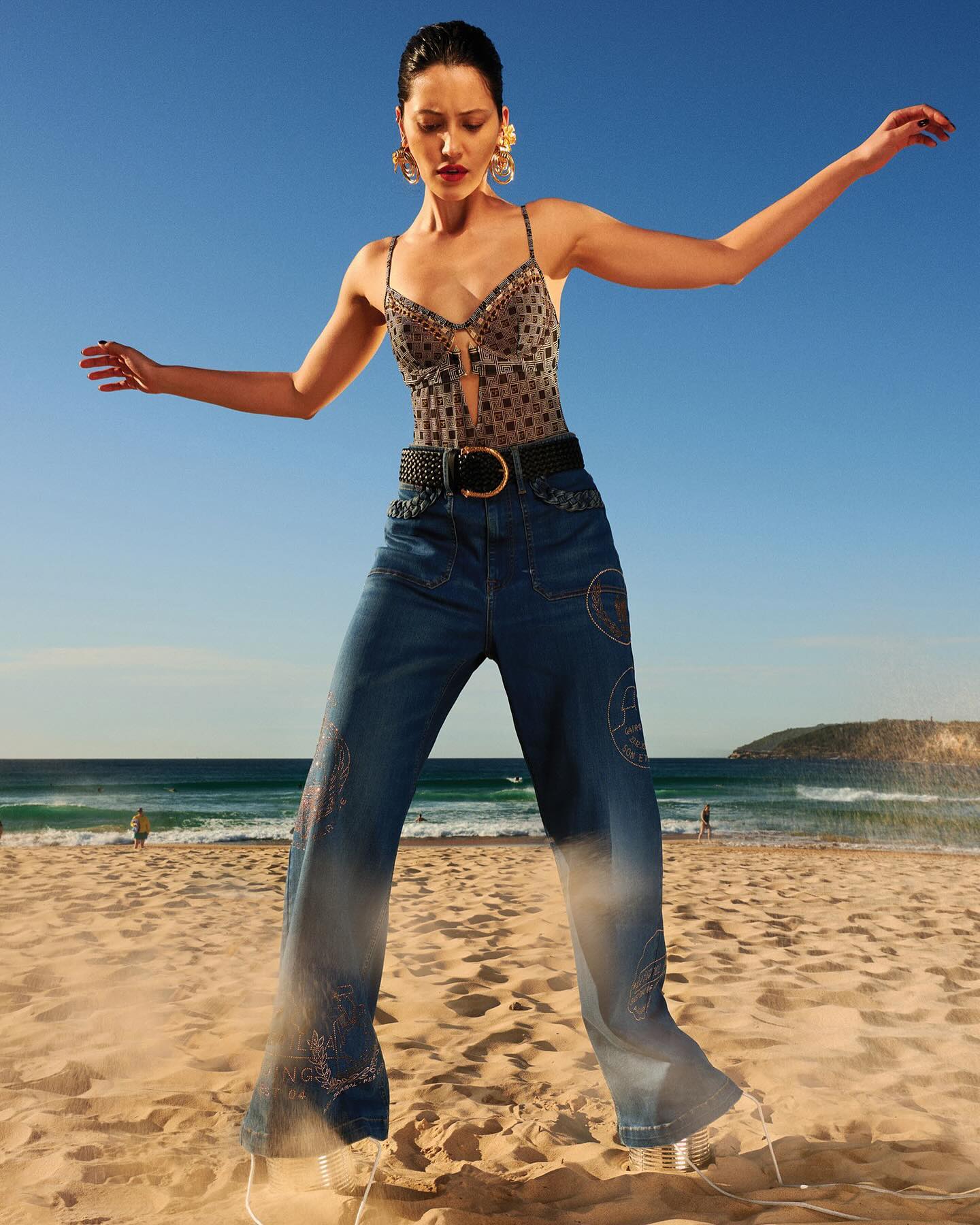 Adult woman in wide-leg jeans and a patterned bodysuit poses on a sandy beach with blue sky, ocean waves, and a distant coastline.