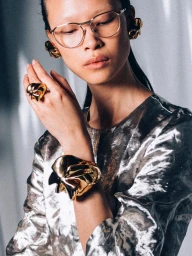Model wearing gold jewellery, wire-rimmed glasses, and a metallic patterned top against a draped fabric backdrop.