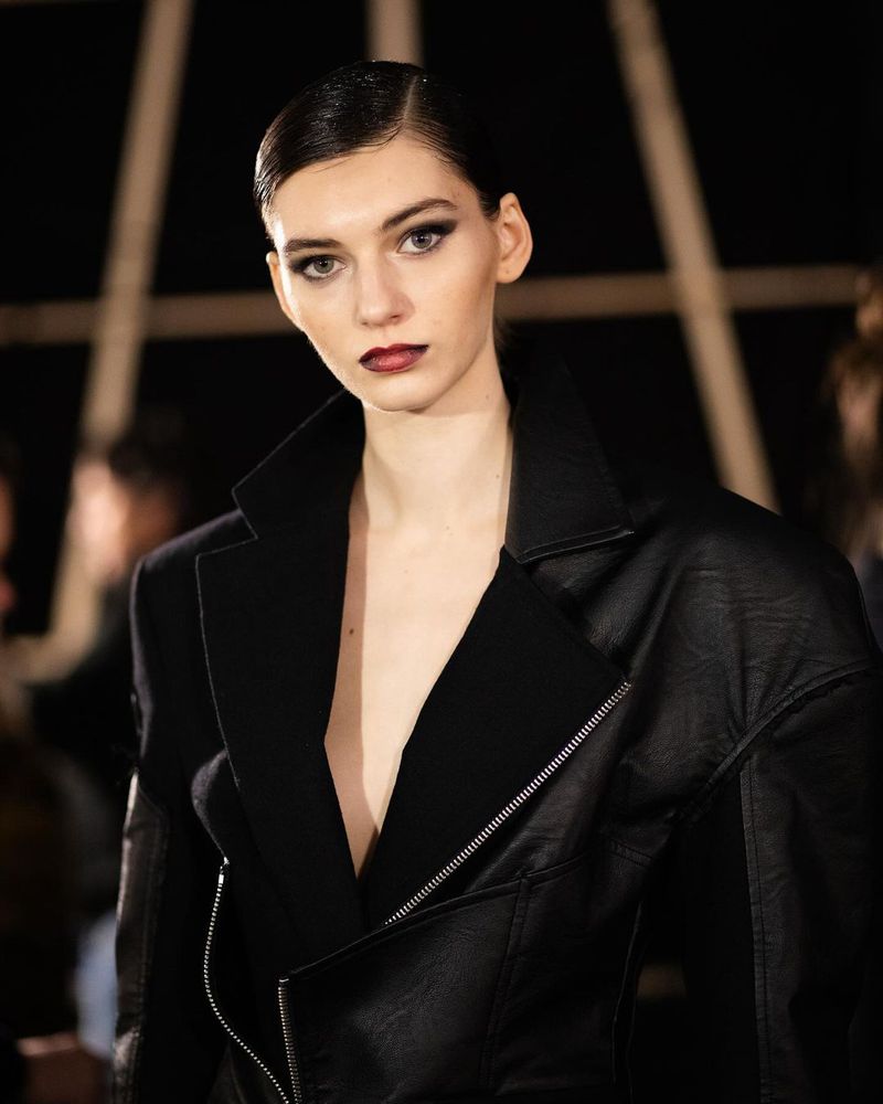 Portrait of a woman with dark hair and makeup wearing a black leather jacket in a dark indoor environment.