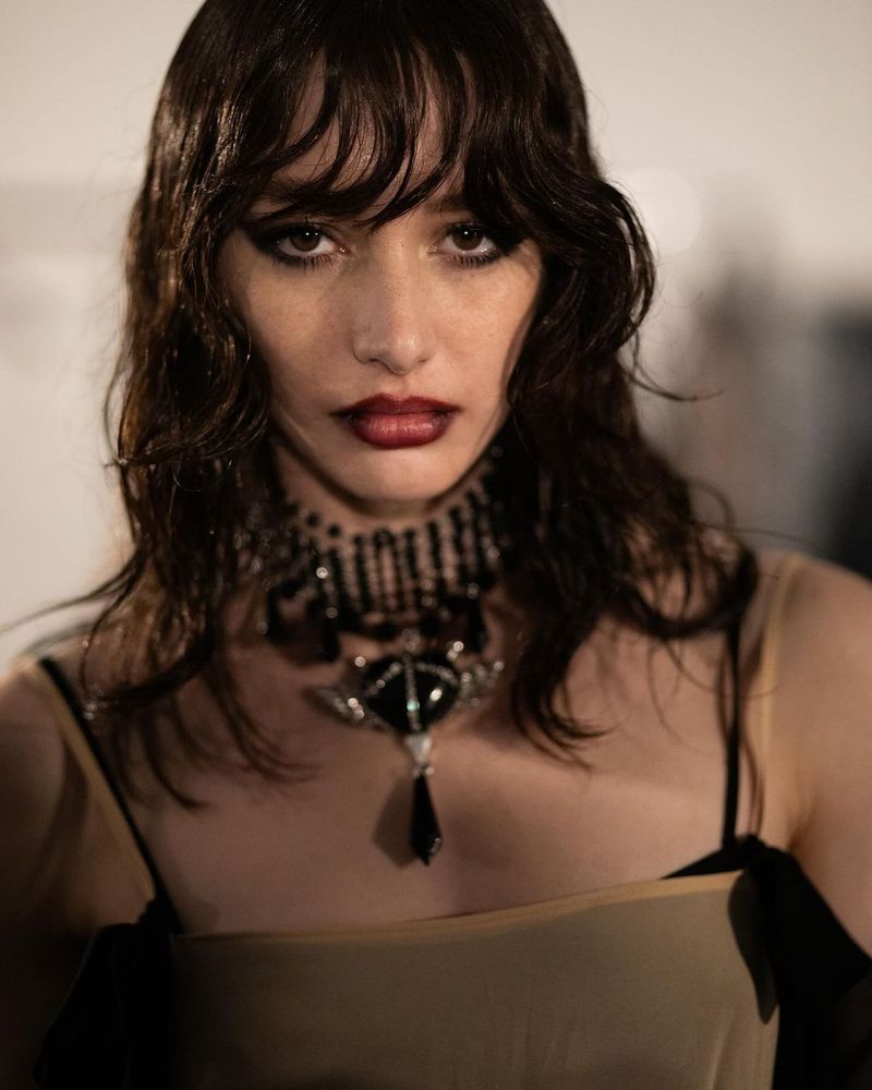 Close-up portrait of a young adult woman with dark hair wearing a beaded choker and dark lipstick against a neutral background.
