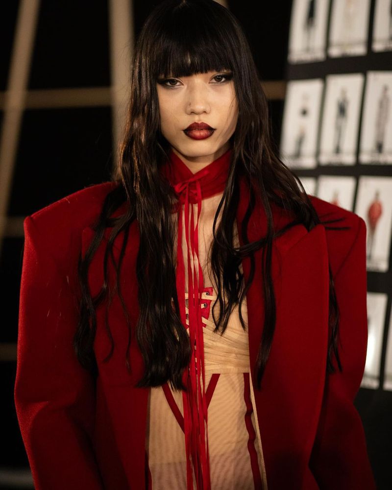 Close-up of an Asian fashion model with dark hair and red lipstick, wearing a red jacket and sheer dress with red accents.