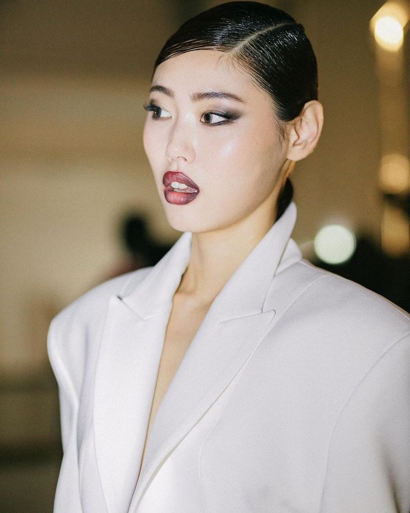Portrait of a fashionable Asian adult female in a white blazer with slicked-back hair and bold makeup, looking to the side in an indoor setting.