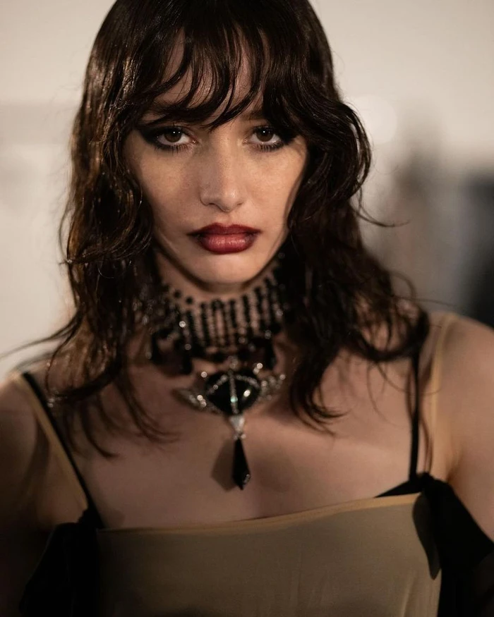 Close-up portrait of a model with dark wavy hair wearing a beaded statement necklace and a dark strap dress.