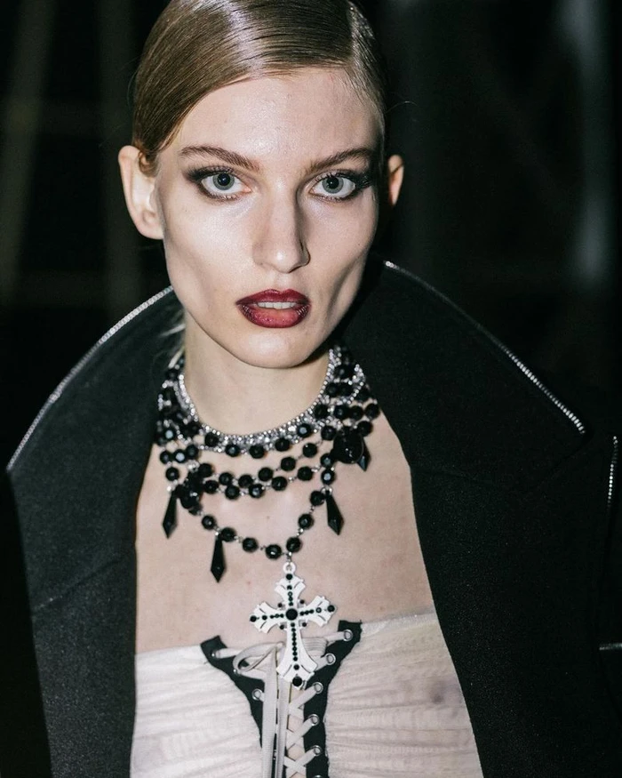Model wearing a layered beaded cross necklace and dark coat in a backstage portrait for Aniye Records Fall Winter 24