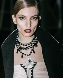 Model wearing a layered beaded cross necklace and dark coat in a backstage portrait for Aniye Records Fall Winter 24