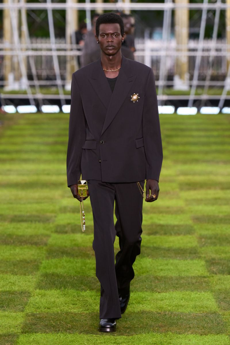 Runway fashion: A slender Black male model strides confidently on a checkered green surface, wearing a dark oversized suit and dress shoes.