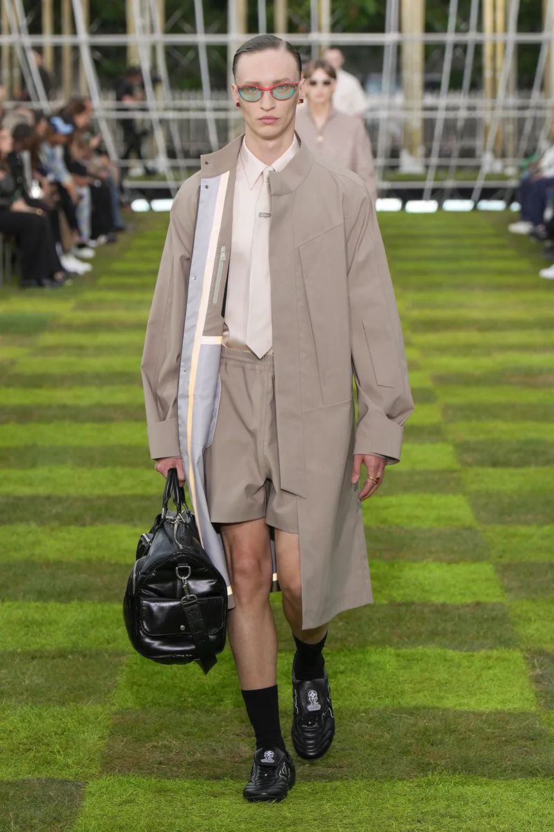 Male model walking on a runway during a fashion show wearing a tan trench coat, shorts, black socks and shoes, and carrying a black bag.