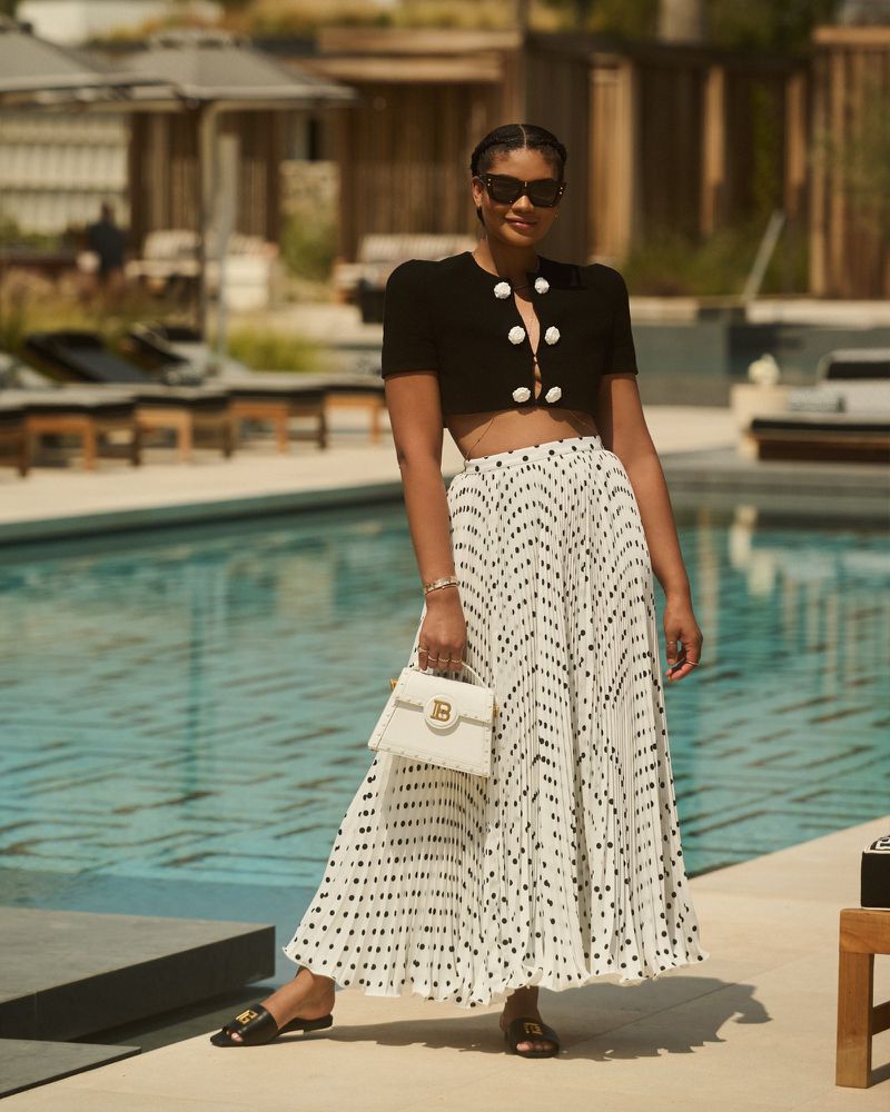 Woman in black cropped top and white polka dot skirt stands by a pool, holding a white handbag in an outdoor setting on a sunny day.