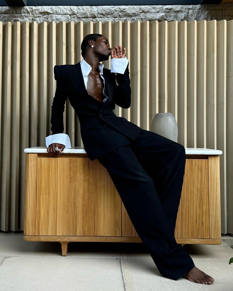 A Black male model in a black suit and white shirt leans against a cabinet against a patterned wall in an indoor setting.