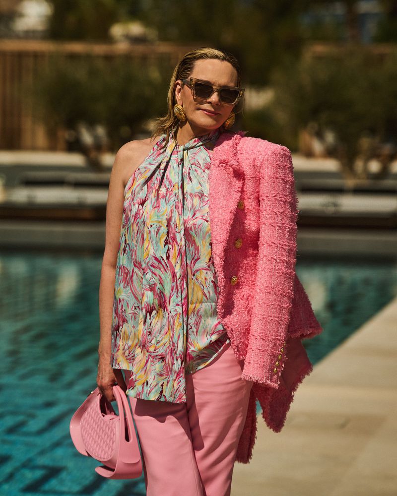 Adult woman in colorful top and pink pants holds pink purse and wears sunglasses and earrings outdoors in natural sunlight.