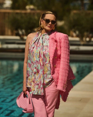 Adult woman in colorful top and pink pants holds pink purse and wears sunglasses and earrings outdoors in natural sunlight.