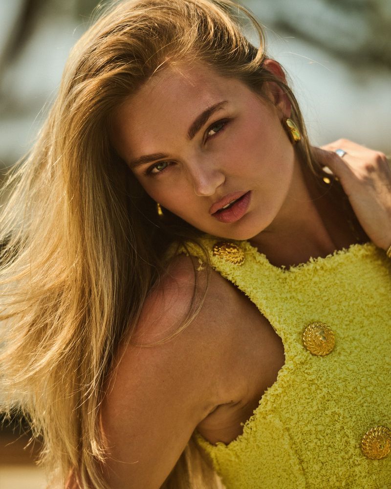 Portrait of a blonde woman with fair skin looking at the camera, wearing a yellow tweed dress with gold buttons and gold earrings.