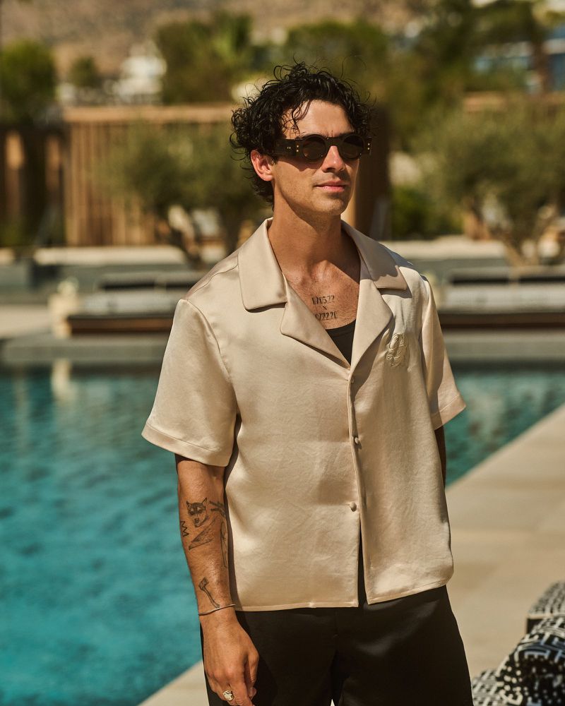 Adult male with curly dark hair wearing sunglasses and a light satin-look shirt poses outdoors near a swimming pool with greenery.