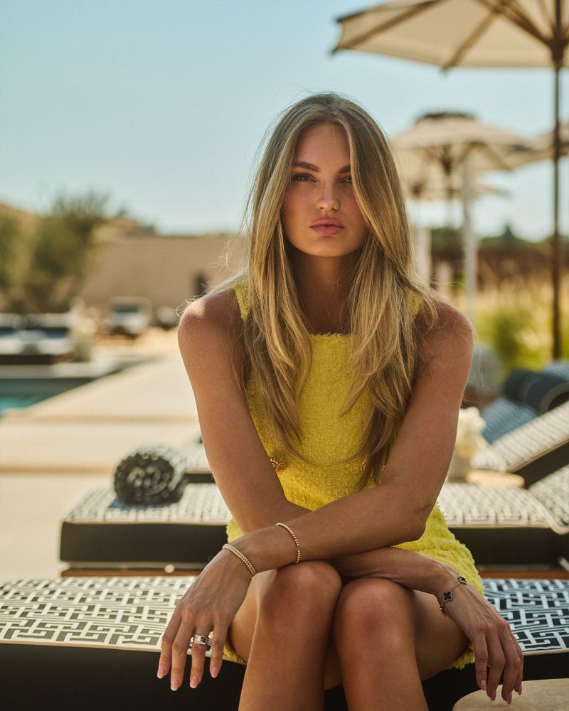 Woman with long blonde hair sits in a yellow dress on a lounge chair by a pool, on a sunny day at an outdoor resort.