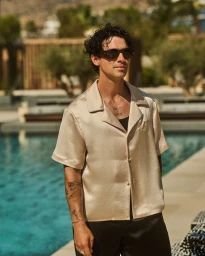 Joe Jonas wearing a satin short-sleeve shirt and dark sunglasses by a swimming pool for Balmain