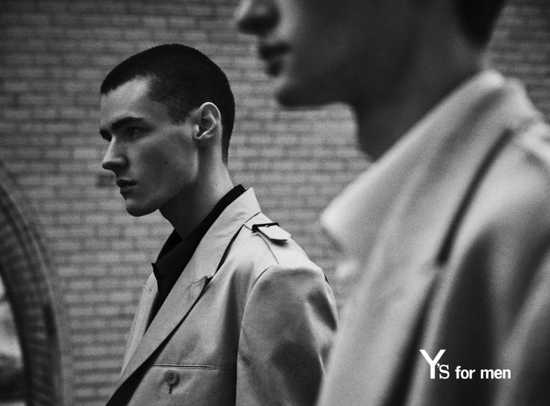 Monochrome shot of two adult men in trench coats against a brick wall, featuring the 'YS for men' logo. Fashion editorial.