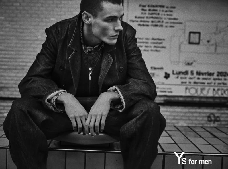 Monochrome fashion shot of a pensive man in a denim jacket and pants, sitting indoors against a textured wall in a timeless style.