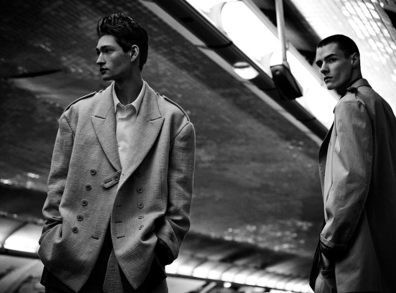 Two adult men in stylish coats stand in an urban setting; a black and white fashion photograph with an industrial background.