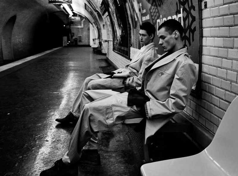 Two men in matching trench coats sit on a bench at a subway station, captured in a monochromatic, editorial fashion style.