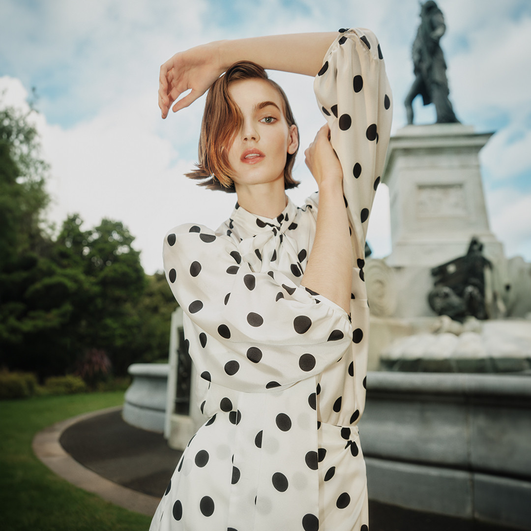 Fashion shot of a young woman in a polka dot dress with a statue and greenery in the background under a cloudy sky.