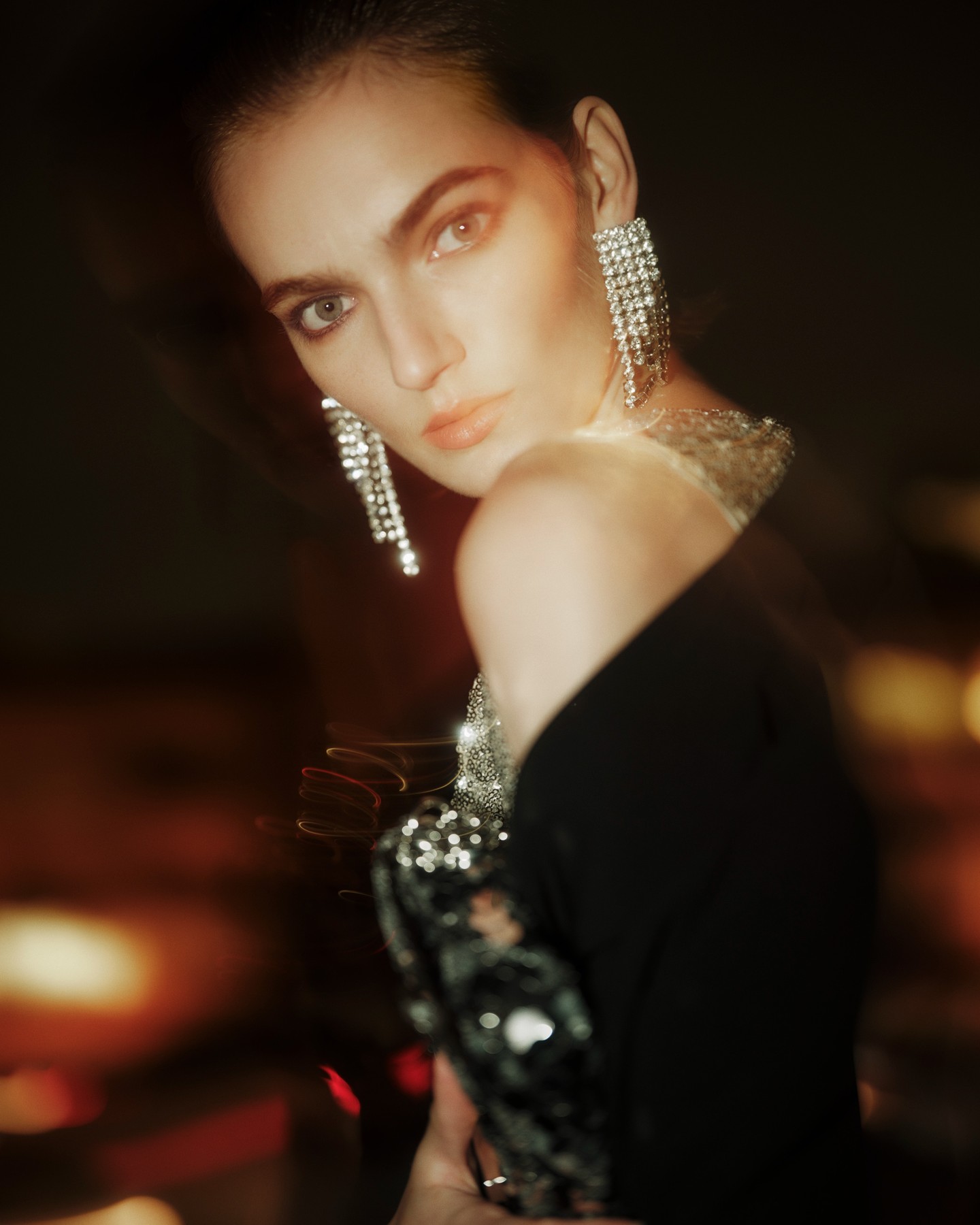 Elegant woman in a black blazer and silver earrings poses in a dimly lit setting with blurred bokeh lights.
