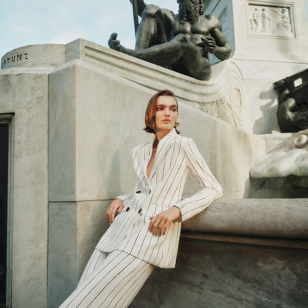 Fashionable woman in a pinstripe suit poses against a stone statue on a sunny day, showcasing a blend of modern style and classic art.