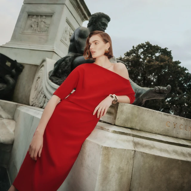 Woman in red dress poses by a statue in an outdoor setting. The medium shot captures the dress's off-the-shoulder design.