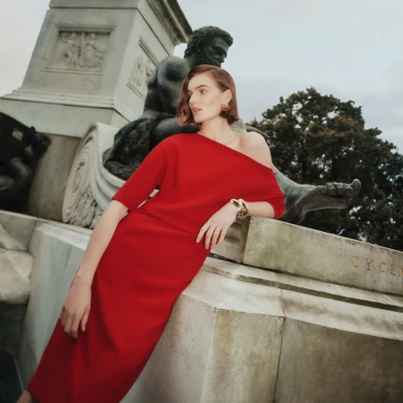 Woman in red dress poses by a statue in an outdoor setting. The medium shot captures the dress's off-the-shoulder design.