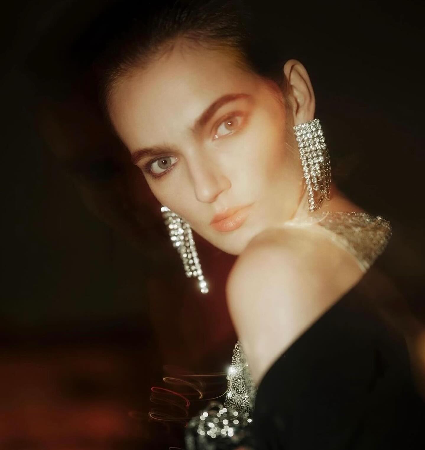 Woman with dramatic silver chandelier earrings wearing a dark dress posing against a blurred dark brown background in a medium shot.