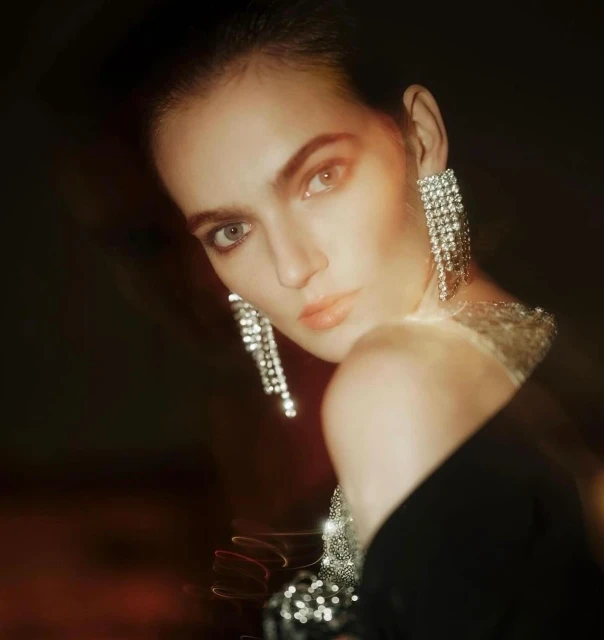 Woman with dramatic silver chandelier earrings wearing a dark dress posing against a blurred dark brown background in a medium shot.