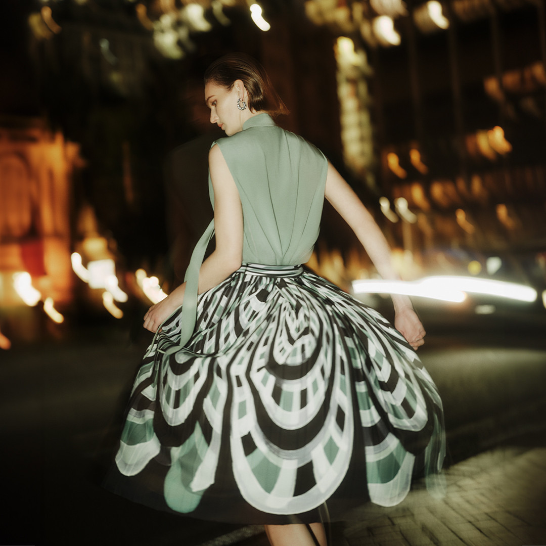 Model in green sleeveless top and patterned flared skirt on a blurred nighttime street for Perri Cutten