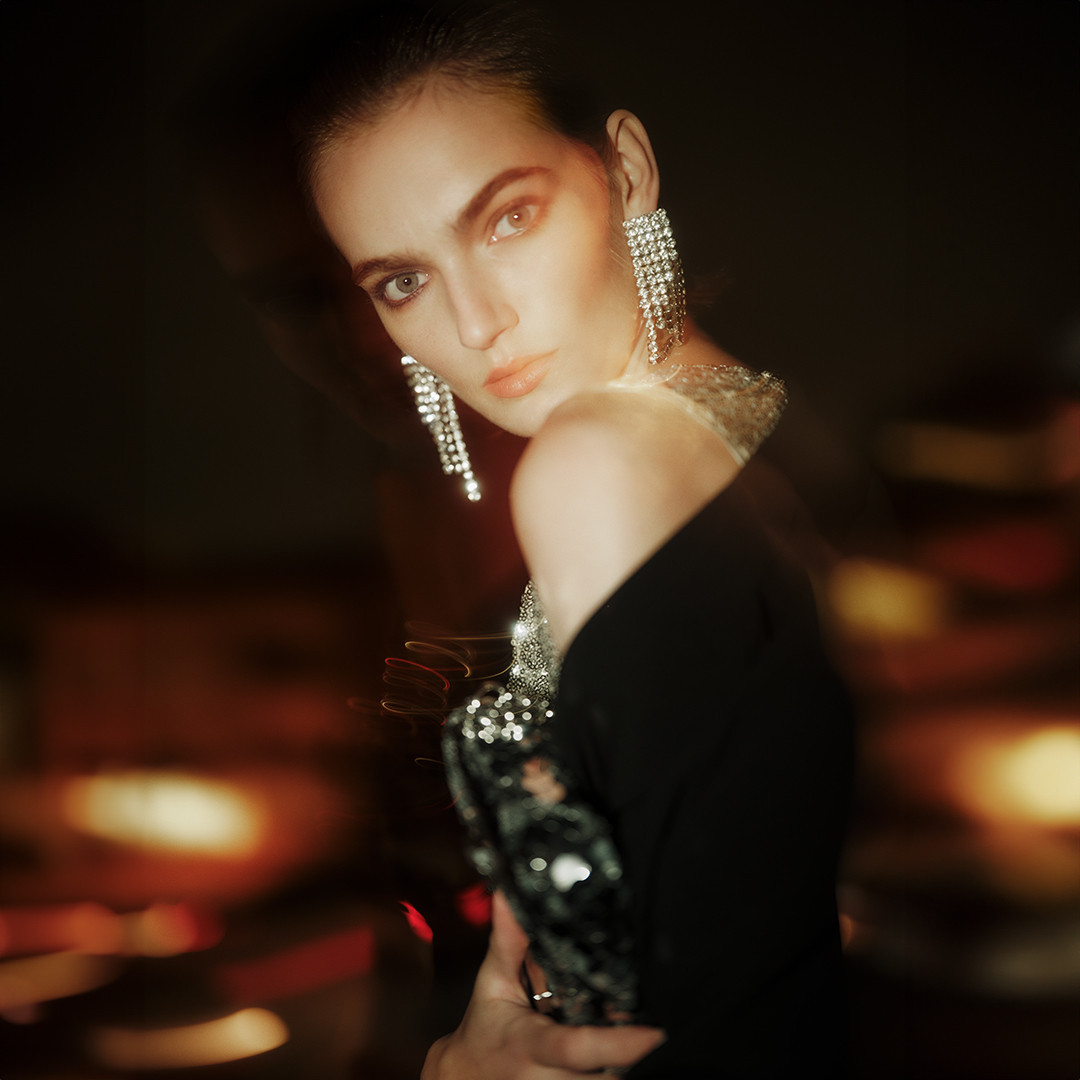 Portrait of an adult female model with dark hair, wearing sparkly earrings, a black dress, and holding a beaded bag in an indoor setting.