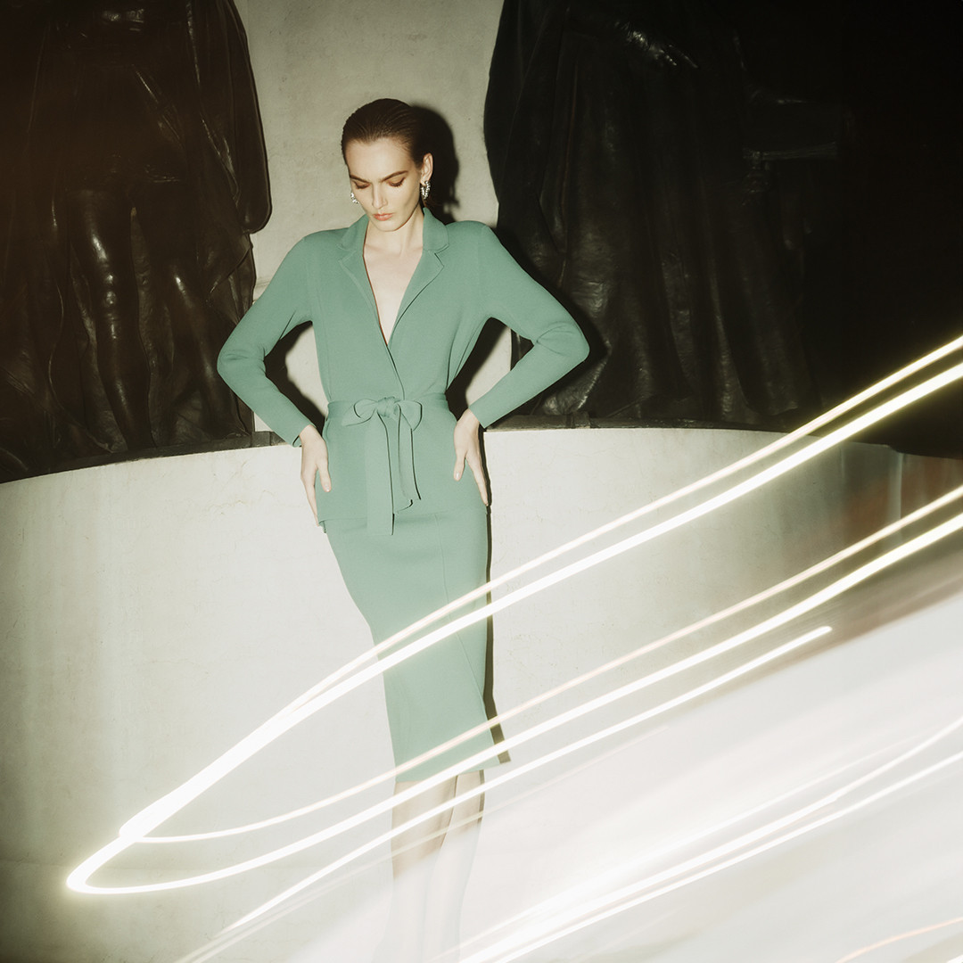 Model wearing a green belted dress standing before dark statues with light trails streaking across the foreground