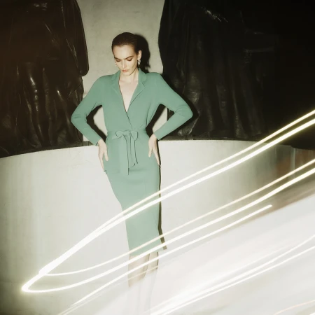 Model wearing a green belted dress standing before dark statues with light trails streaking across the foreground