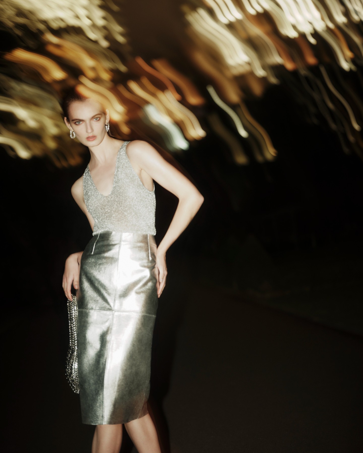 Fashion shot of a female model wearing a silver top and skirt, holding a silver purse, posing in front of a dark background.