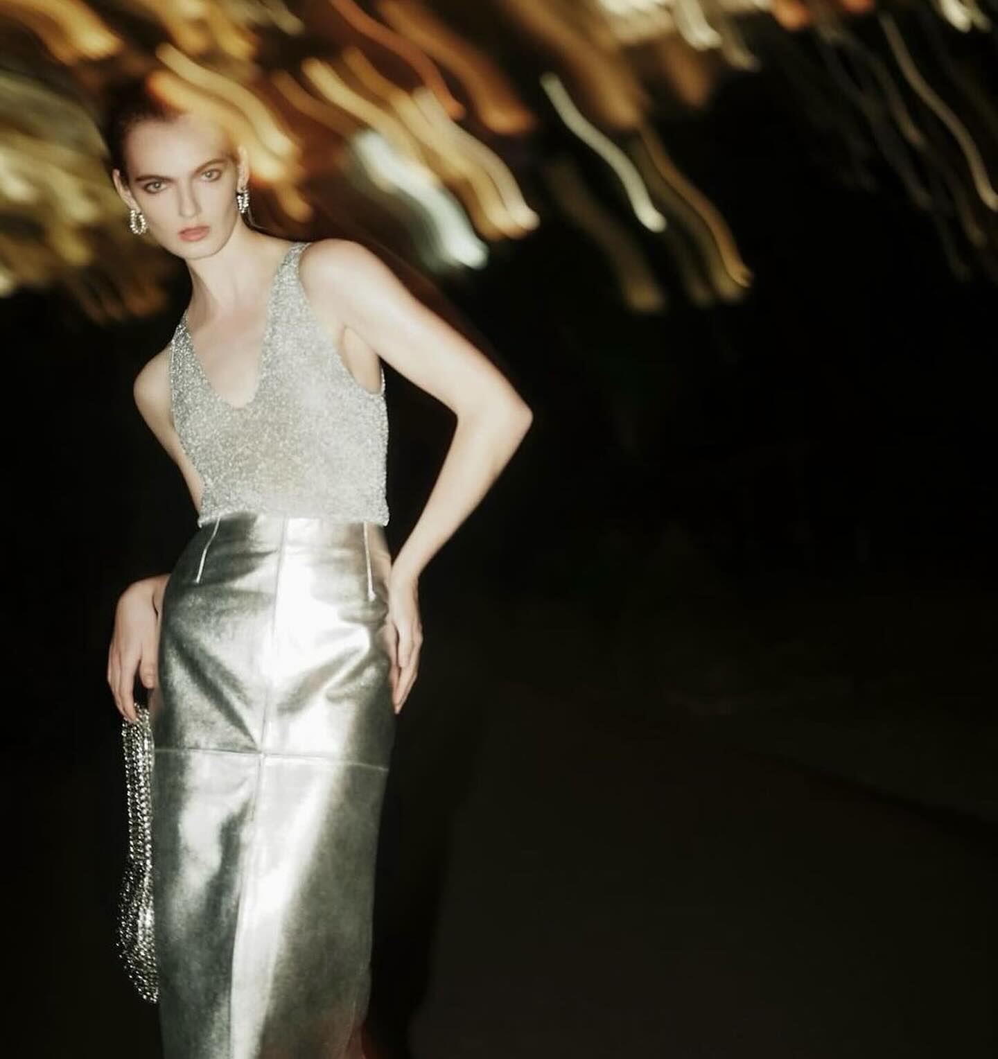 A slender fashion model poses with hands on hips, wearing a silver beaded top and skirt against a dark background with blurred lights.