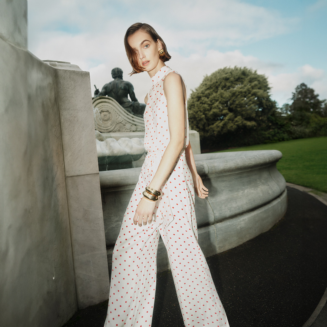 A fashionable adult woman is the focus, wearing a white jumpsuit with red dots, accessorized with gold earrings and bracelets, standing outdoors.