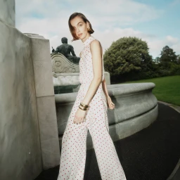 Model in white polka dot jumpsuit walking near stone fountain and park greenery for SS24 fashion project