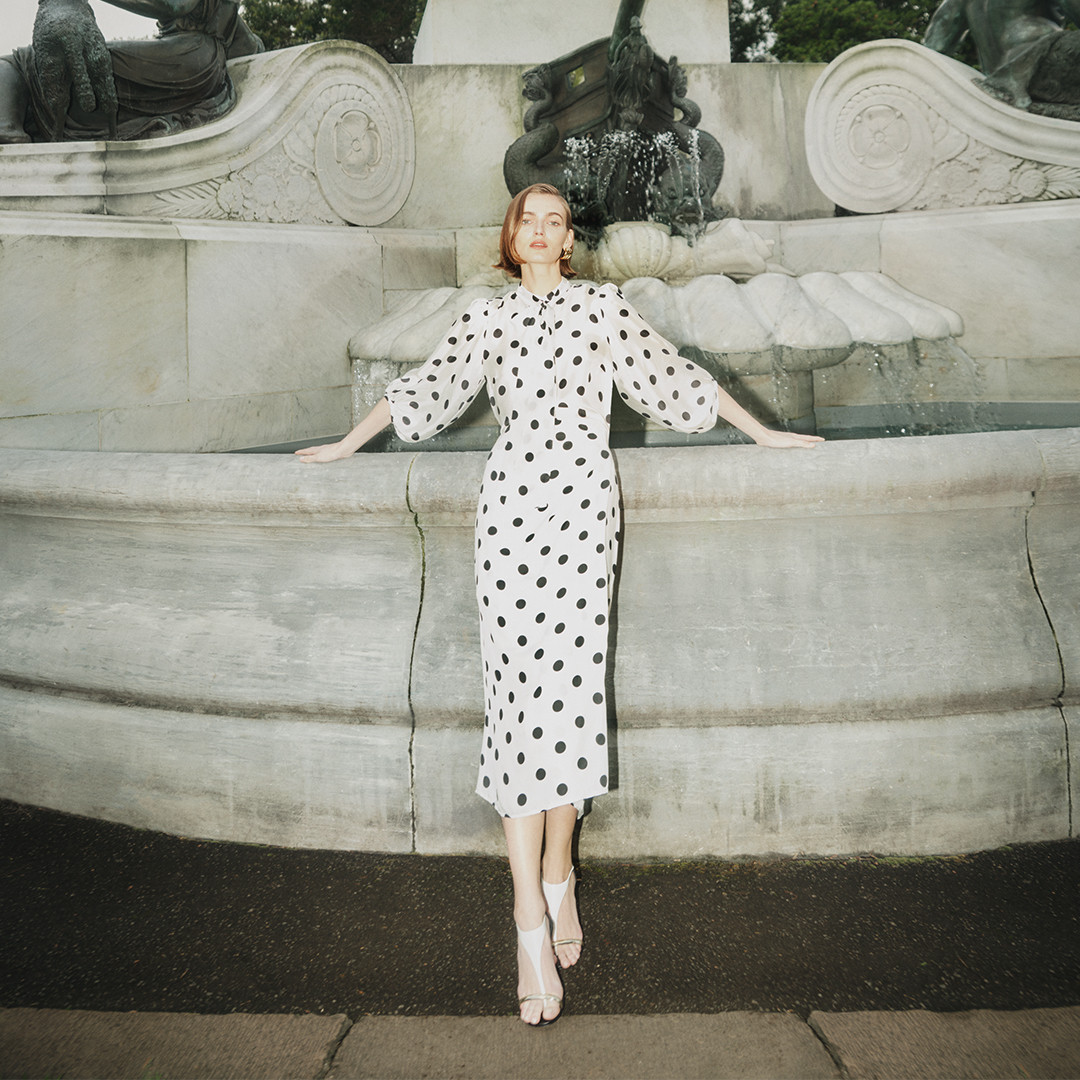 A woman in a polka dot dress poses near a stone fountain, wearing white high-heeled sandals, under daylight in an outdoor setting.