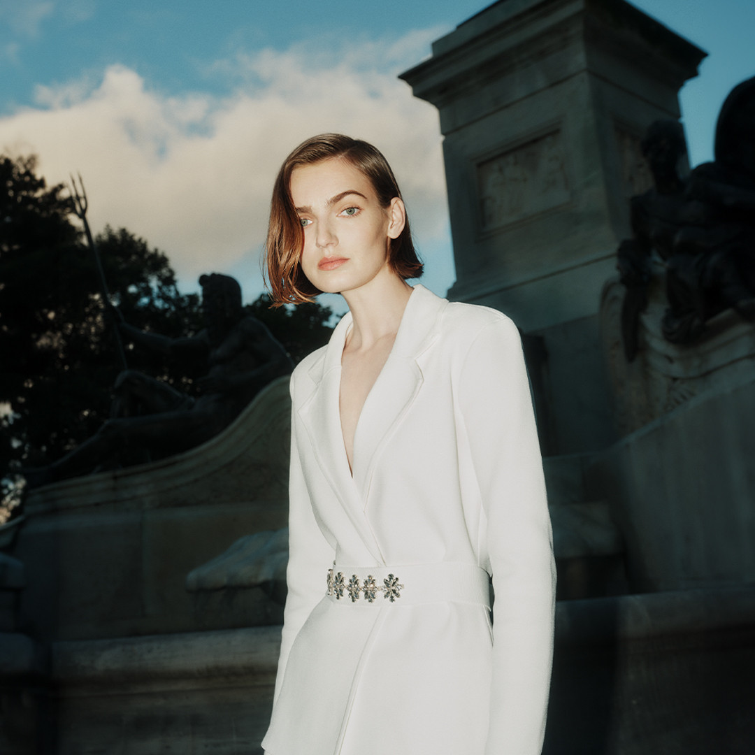 Elegant woman in a white dress and blazer stands confidently in front of a monument with a cloudy sky in the background.