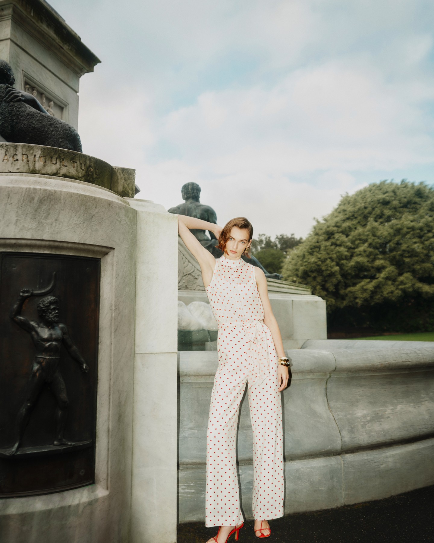An adult woman poses in a sleeveless polka dot jumpsuit and red heels in a park setting next to a decorative stone monument.