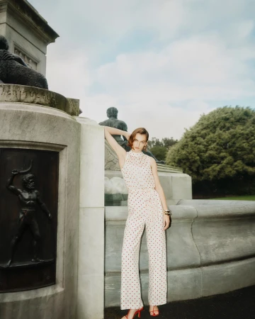An adult woman poses in a sleeveless polka dot jumpsuit and red heels in a park setting next to a decorative stone monument.