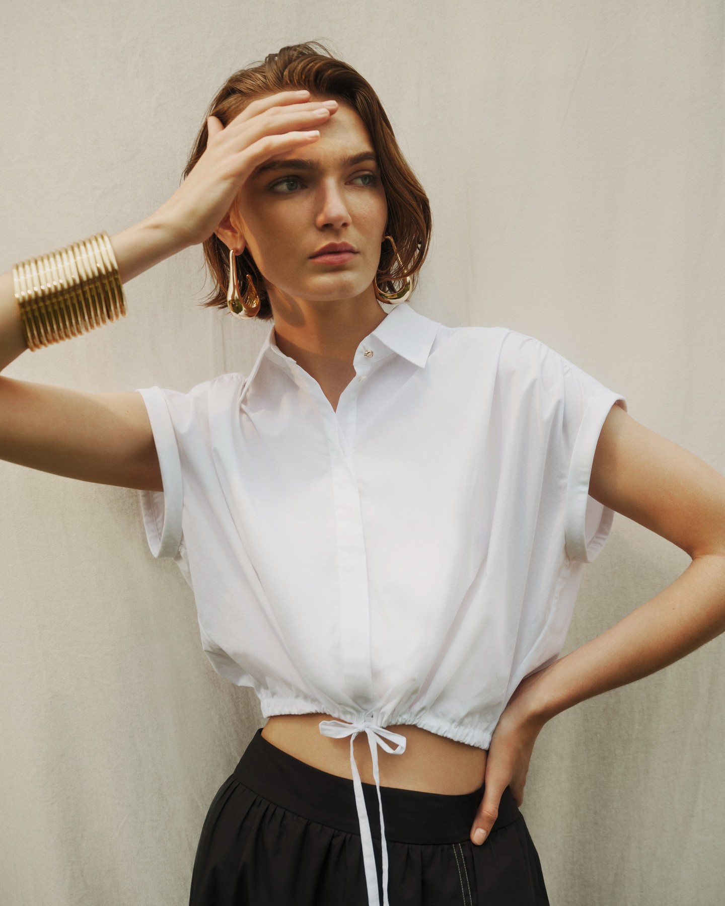 Model wearing a white crop shirt with a drawstring hem and large gold bangles against a neutral background.