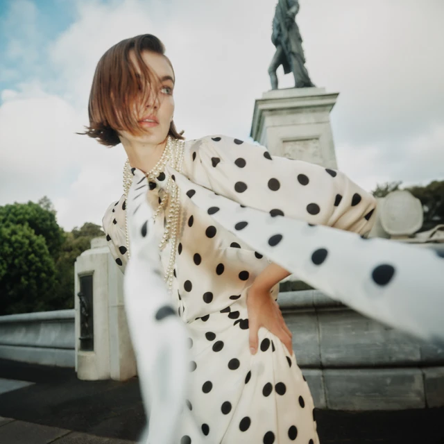 Woman in polka dot dress with pearl necklace poses near stone monument outdoors under cloudy sky, fashion editorial photography.