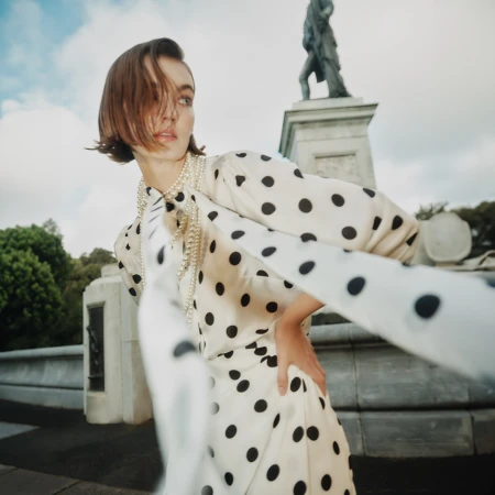 Woman in polka dot dress with pearl necklace poses near stone monument outdoors under cloudy sky, fashion editorial photography.