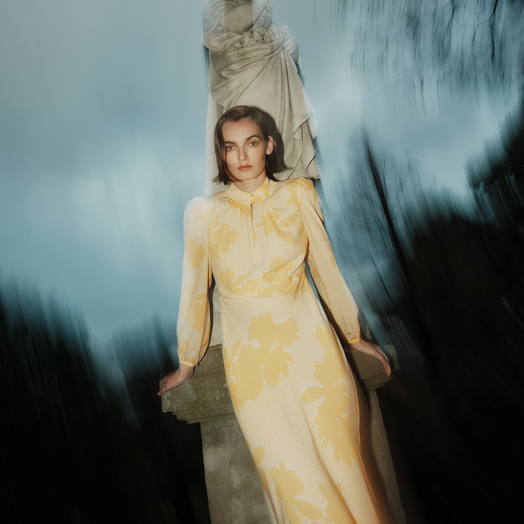 Adult woman in a yellow floral dress poses next to a stone sculpture against a blurred background with trees and cloudy skies.