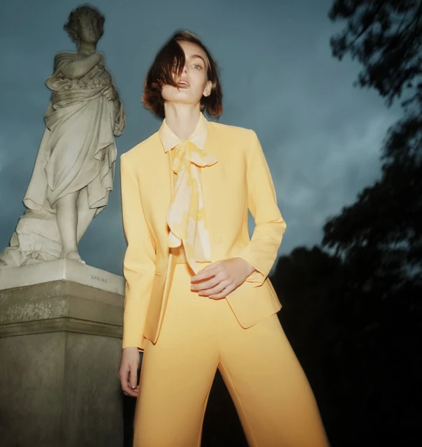 Woman poses in yellow pantsuit by marble statue in an outdoor scene with dramatic sky, fashion-forward and classic style.