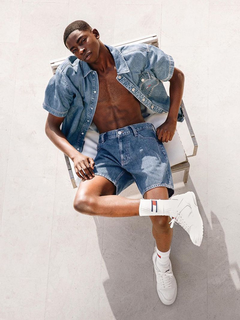A young adult male model lies on a lounge chair wearing denim shorts, an unbuttoned short-sleeve denim shirt, socks, and sneakers.