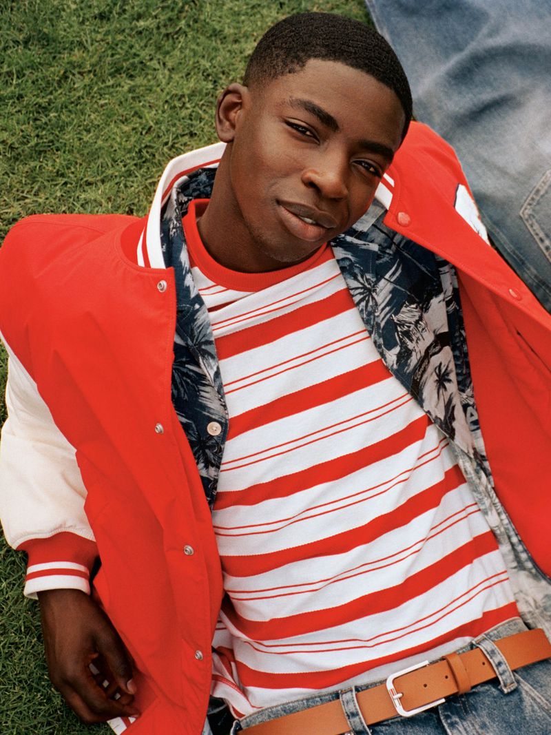 Young Black man in an open varsity jacket and striped shirt poses on a green lawn with denim-clad legs in the background.